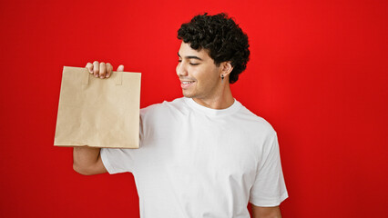 Young latin man smiling confident holding paper bag over isolated red background