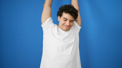 Young latin man stretching arms over isolated blue background - obrazy, fototapety, plakaty