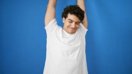 Foto op Canvas Young latin man stretching arms over isolated blue background © Krakenimages.com