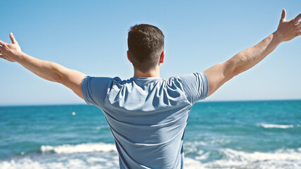 Young hispanic man tourist standing with arms open on back view at seaside