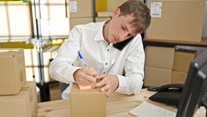 Young caucasian man ecommerce business worker writing on package talking on smartphone at office