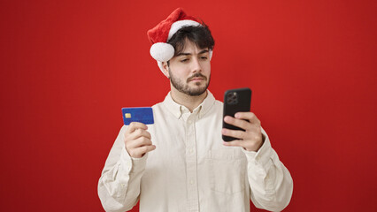 Young hispanic man using smartphone and credit card wearing christmas hat over isolated red background