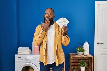 African american man holding money at laundry room covering mouth with hand, shocked and afraid for...