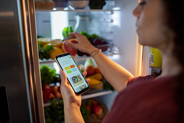 Mixed middle aged woman using her diet app on her smartphone while looking through her fridge at home