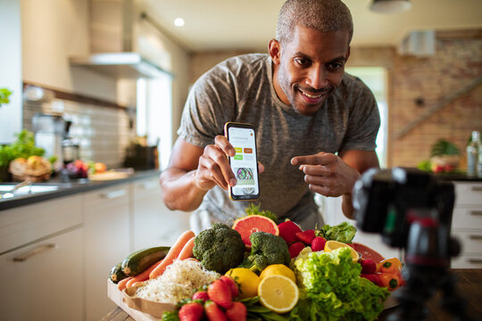 Middle aged african american male food nutritionist and blogger promoting and showing his dieting app to his customers and clients while recording in the kitchen with organic fruits and vegetables