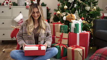 Young blonde woman unpacking gift sitting by christmas tree at home