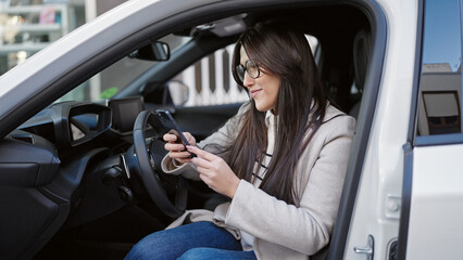 Young beautiful hispanic woman using smartphone sitting on car at street