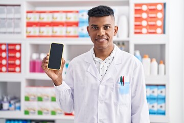 Young hispanic man working at pharmacy drugstore showing smartphone screen looking positive and happy standing and smiling with a confident smile showing teeth