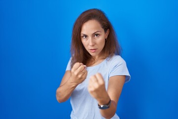 Brunette woman standing over blue background ready to fight with fist defense gesture, angry and...