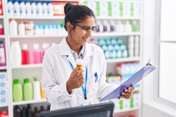 Young beautiful hispanic woman pharmacist holding pills bottle reading document at pharmacy