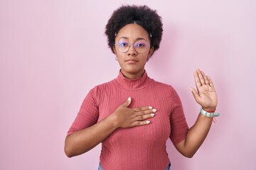 Beautiful african woman with curly hair standing over pink background swearing with hand on chest...