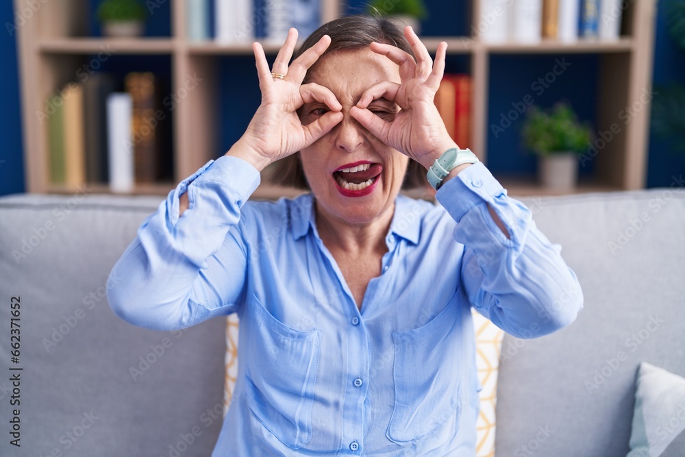 Canvas Prints Middle age hispanic woman sitting on the sofa at home doing ok gesture like binoculars sticking tongue out, eyes looking through fingers. crazy expression.