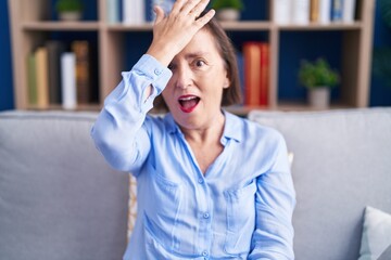 Middle age hispanic woman sitting on the sofa at home surprised with hand on head for mistake,...