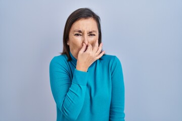 Middle age hispanic woman standing over isolated background smelling something stinky and disgusting, intolerable smell, holding breath with fingers on nose. bad smell