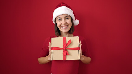Young beautiful hispanic woman wearing christmas hat holding gift over isolated red background