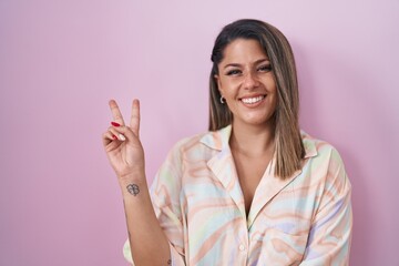 Blonde woman standing over pink background smiling with happy face winking at the camera doing victory sign. number two.