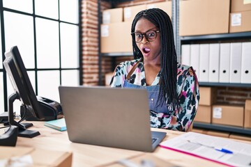 African woman with braids working at small business ecommerce with laptop scared and amazed with open mouth for surprise, disbelief face - obrazy, fototapety, plakaty
