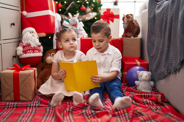 Obraz na płótnie Canvas Two kids reading book sitting on floor by christmas tree at home
