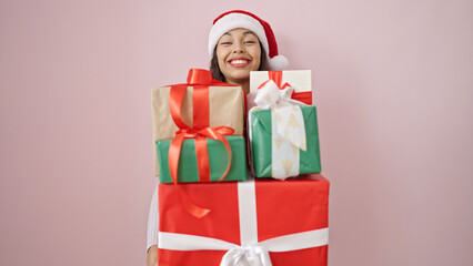 Young beautiful hispanic woman wearing christmas hat holding gifts over isolated pink background