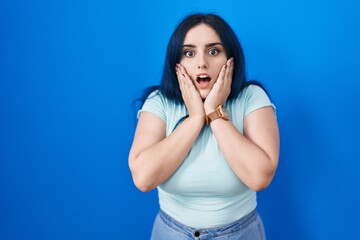 Young modern girl with blue hair standing over blue background afraid and shocked, surprise and amazed expression with hands on face