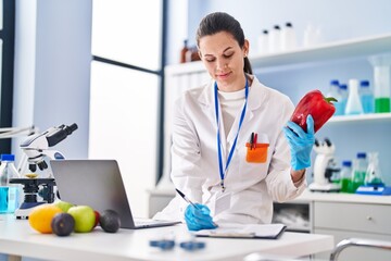 Young beautiful hispanic woman scientist holding pepper writing on document at laboratory