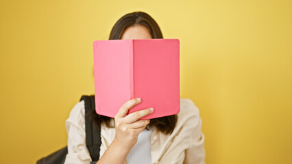 Feisty young hispanic girl student, in casual wear, nonchalantly covering her face with a book against a pop of isolated yellow background wall. the lively photo captures her school life.