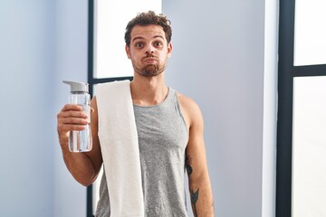 Young hispanic man wearing sportswear drinking water puffing cheeks with funny face. mouth inflated...