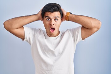 Hispanic man standing over blue background crazy and scared with hands on head, afraid and surprised of shock with open mouth