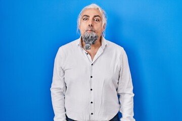 Middle age man with grey hair standing over blue background making fish face with lips, crazy and comical gesture. funny expression.