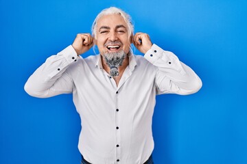 Middle age man with grey hair standing over blue background smiling pulling ears with fingers, funny gesture. audition problem