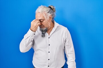 Middle age man with grey hair standing over blue background tired rubbing nose and eyes feeling fatigue and headache. stress and frustration concept.