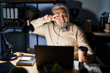 Middle age man with grey hair working at the office at night doing peace symbol with fingers over face, smiling cheerful showing victory