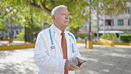 Senior grey-haired man doctor standing with serious expression using smartphone at park