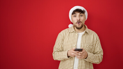 Young arab man wearing christmas hat using smartphone over isolated red background