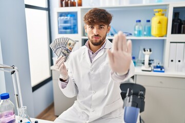 Arab man with beard working at scientist laboratory holding money with open hand doing stop sign with serious and confident expression, defense gesture