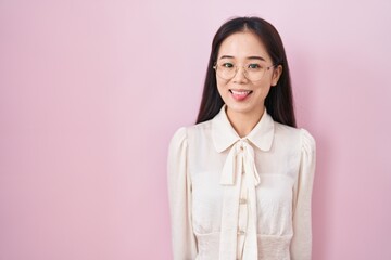 Young chinese woman standing over pink background sticking tongue out happy with funny expression....