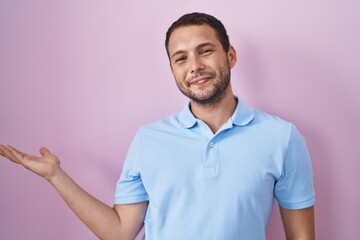 Fototapeta premium Hispanic man standing over pink background smiling cheerful presenting and pointing with palm of hand looking at the camera.