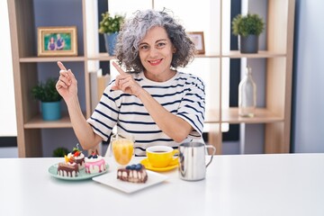 Middle age woman with grey hair eating pastries and drinking coffee for breakfast smiling and looking at the camera pointing with two hands and fingers to the side.