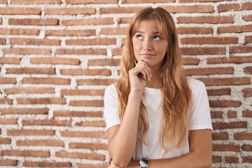 Young caucasian woman standing over bricks wall with hand on chin thinking about question, pensive expression. smiling with thoughtful face. doubt concept.