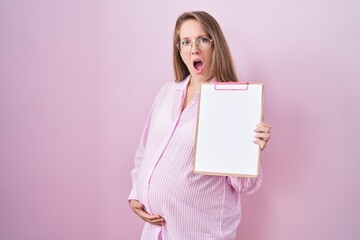 Fototapeta na wymiar Young pregnant woman holding clipboard in shock face, looking skeptical and sarcastic, surprised with open mouth