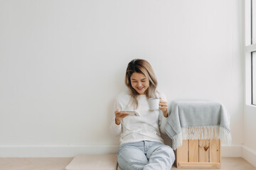 Asian Thai woman enjoy using and holding smartphone in apartment winter time. Happy moment smiling and chilling alone, sitting on floor, drink coffee.
