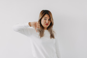 Asian Thai woman grumpy face, showing thumbs down, disapproved sign gesture, dislike prohibit action, giving negative feedback, rejecting no, isolated on white background in winter. Body language.