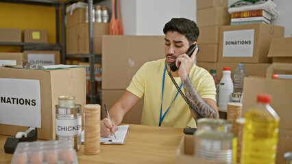 Young hispanic man volunteer talking on telephone writing on clipboard at charity center