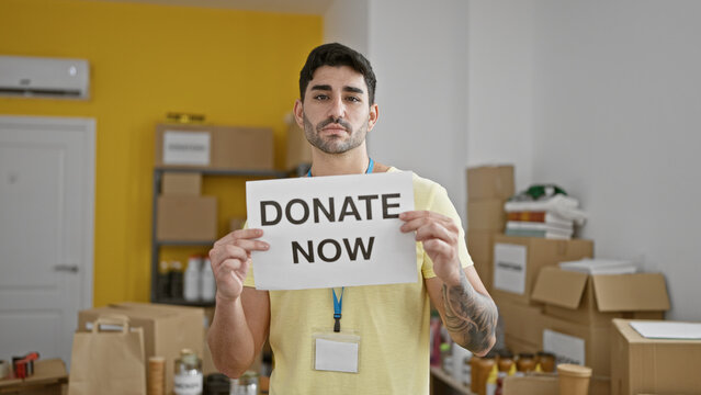 Young hispanic man volunteer holding donate now banner with serious face at charity center