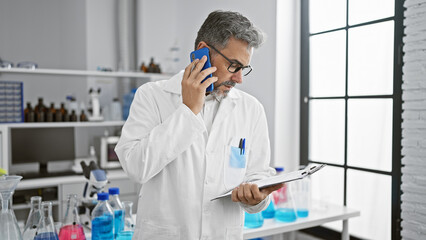 Young, handsome, grey-haired hispanic man, a scientist mastering technology, engrossed in reading documents and talking over his smartphone while working intently at his laboratory table.