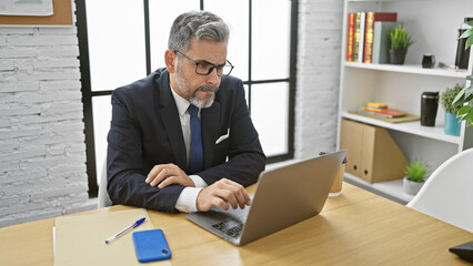 Attractive, grey-haired young hispanic man, an elegant professional, concentrating hard in the office. he's working seriously, managing business success from his laptop at his desk.