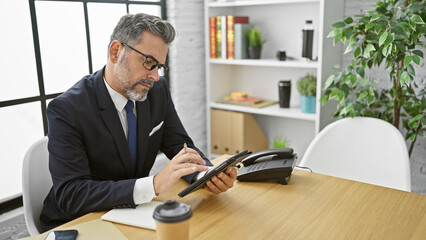 Serious young hispanic man, grey-haired and elegant, deeply engrossed in thought at his workplace while skillfully maneuvering touchpad. indoor office scene captures him in professional attire. - obrazy, fototapety, plakaty