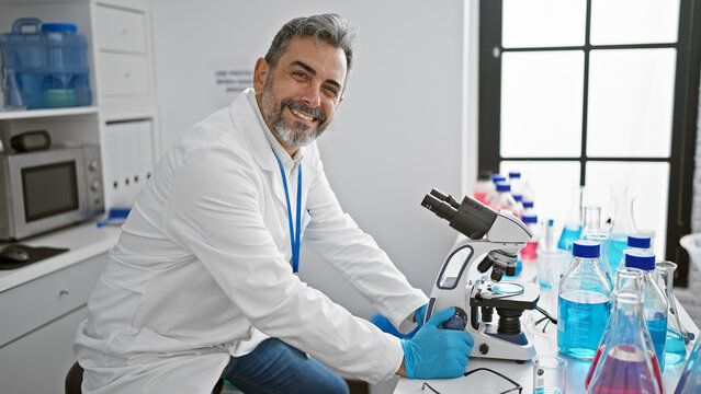 Confident young hispanic scientist with grey hair, smiling as he works with a microscope in the lab, making medical discoveries