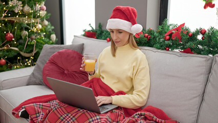 Young blonde woman using laptop drinking orange juice sitting by christmas tree at home