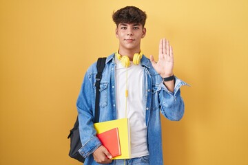 Hispanic teenager wearing student backpack and holding books waiving saying hello happy and...